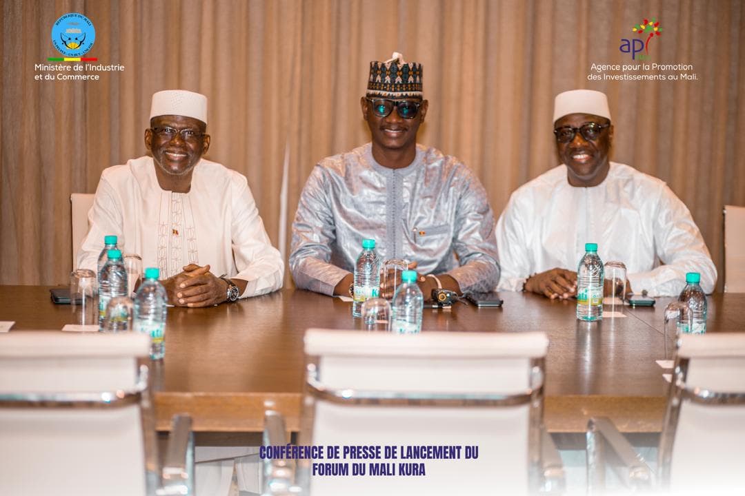 Three men in boubous, two wearing white caps and one an embroidered hat, seated side by side and smiling at a meeting table set with water bottles, with the logos of the Ministry of Industry and Trade and of API-Mali at the top of the frame. — API-Mali