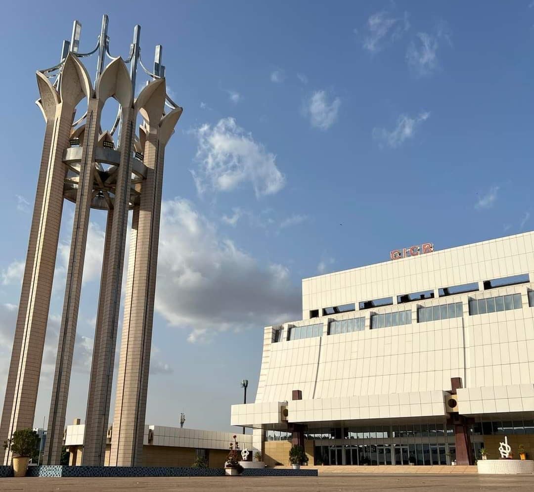 Low-angle view of the Bamako International Conference Centre from its forecourt: a white tile-clad building whose upper storey cantilevers over a sand-coloured stone base, an orange CICB sign on the roof line, the glazed ground-floor entrance behind square columns, potted plants and small sculptures on the paved forecourt, and at left a tall monument of four concrete pillars carrying white petal-shaped forms.