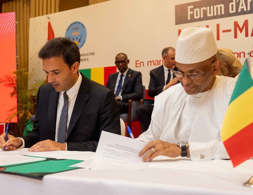 An agreement being signed: a man in a dark suit and a man in a white grand boubou and white cap sign documents side by side at a white-clothed table, a Malian flag in the foreground, and two officials seated behind them in front of a business-forum banner.