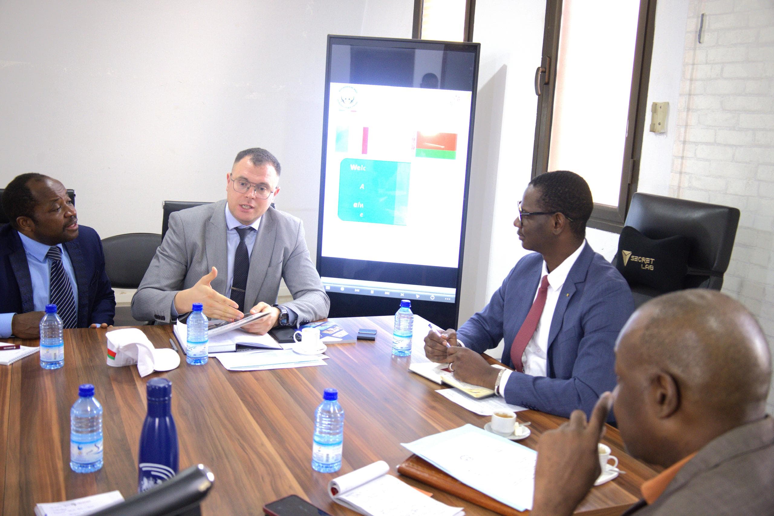 Quatre hommes en costume assis autour d’une table de réunion en bois, en discussion, un écran mural affichant une diapositive d’accueil derrière eux. — API-Mali