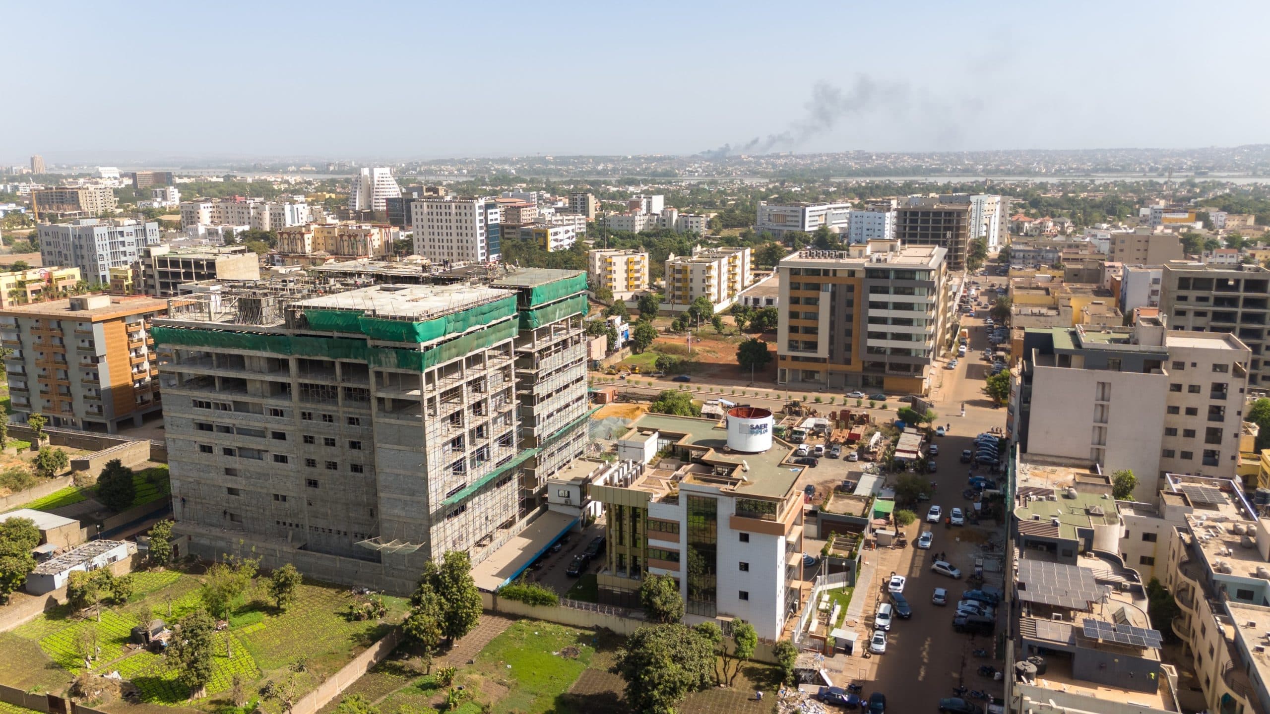 Aerial view of central Bamako: a large concrete tower under construction among mid-rise blocks, with the river and hills on the horizon. — API-Mali