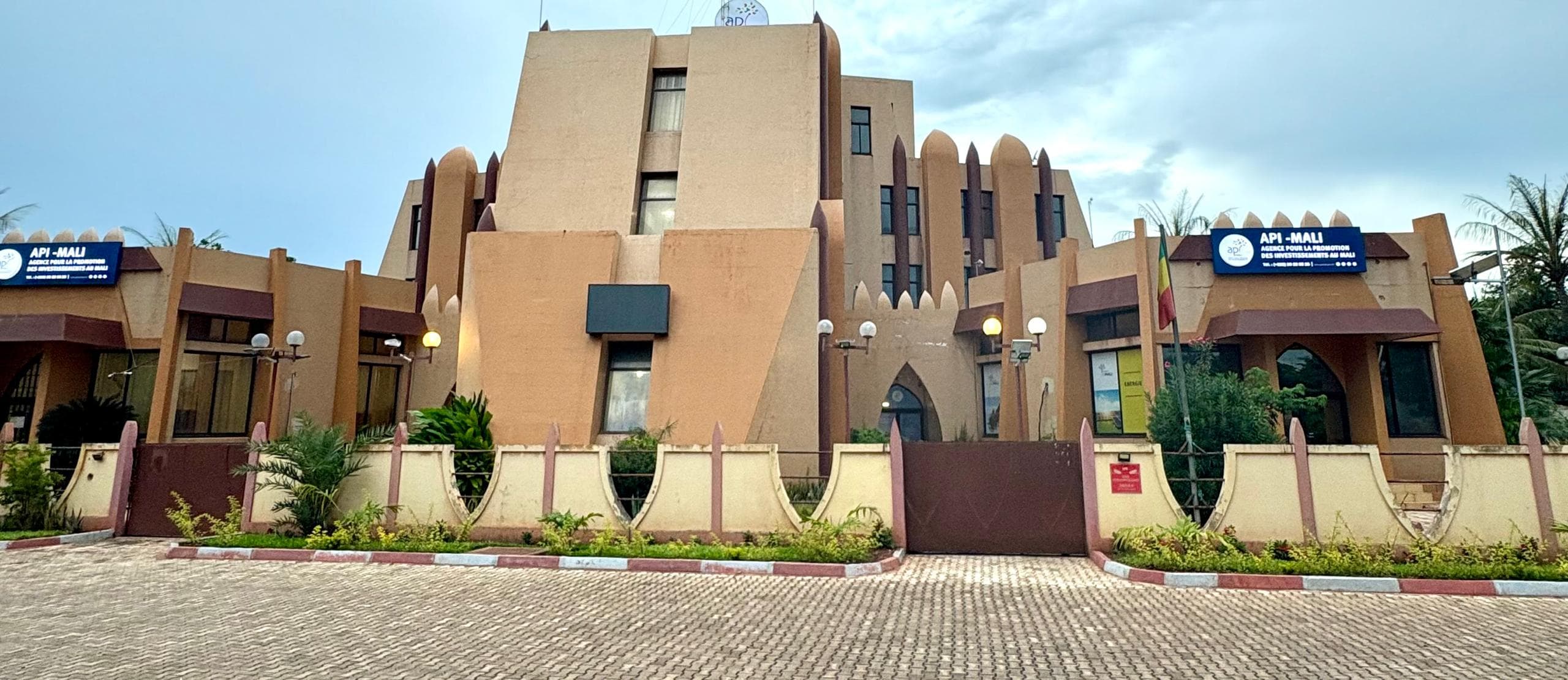 Façade du siège de l’API-Mali à Bamako : bâtiment beige de style soudano-sahélien à pilastres bordeaux, deux panneaux « API-MALI », un drapeau malien et un portail fermé. — API-Mali