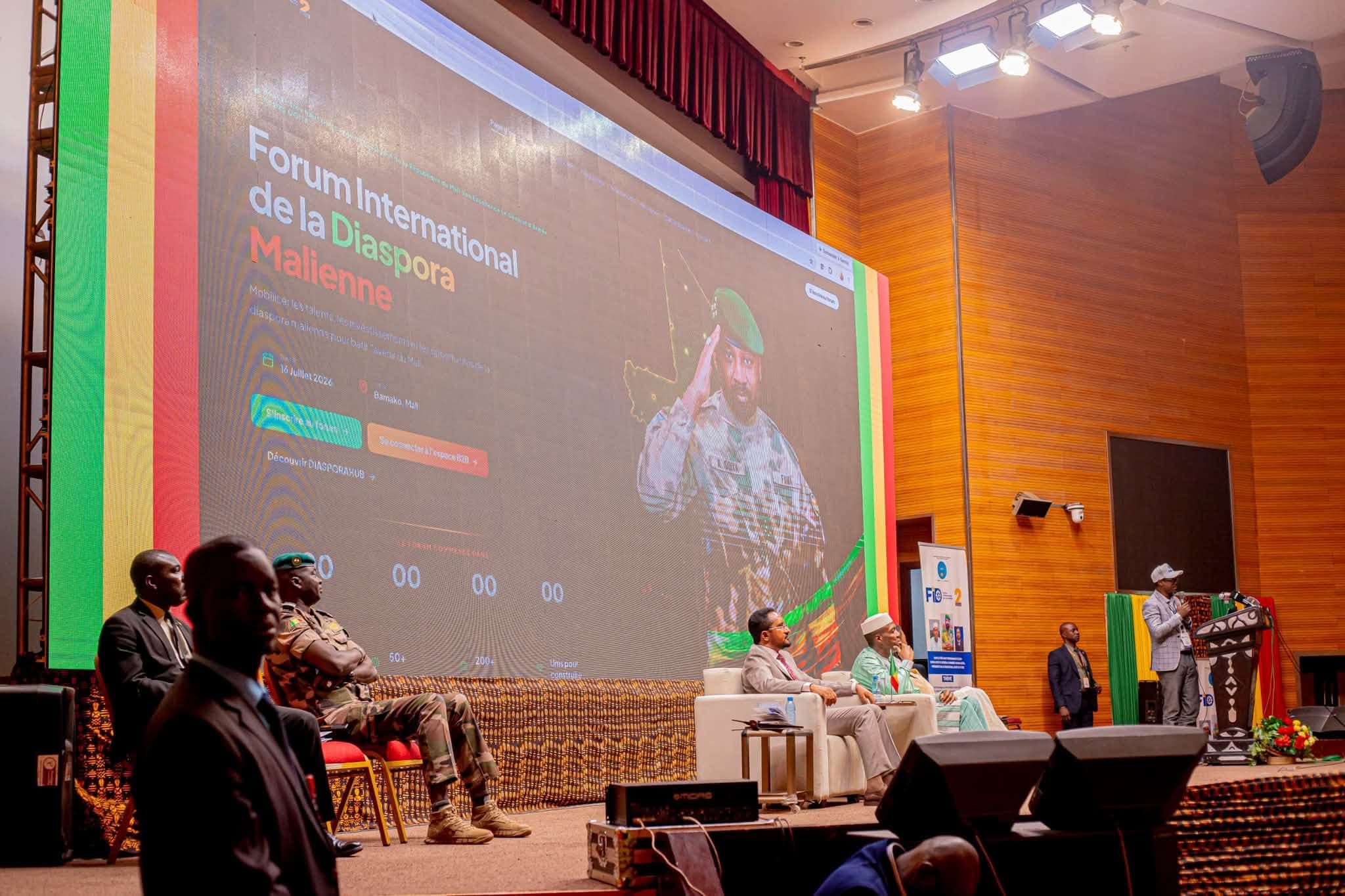 Scène d’un vaste auditorium lambrissé avec un écran LED géant bordé aux couleurs du drapeau malien, deux panélistes en fauteuils blancs et un orateur au pupitre. — API-Mali