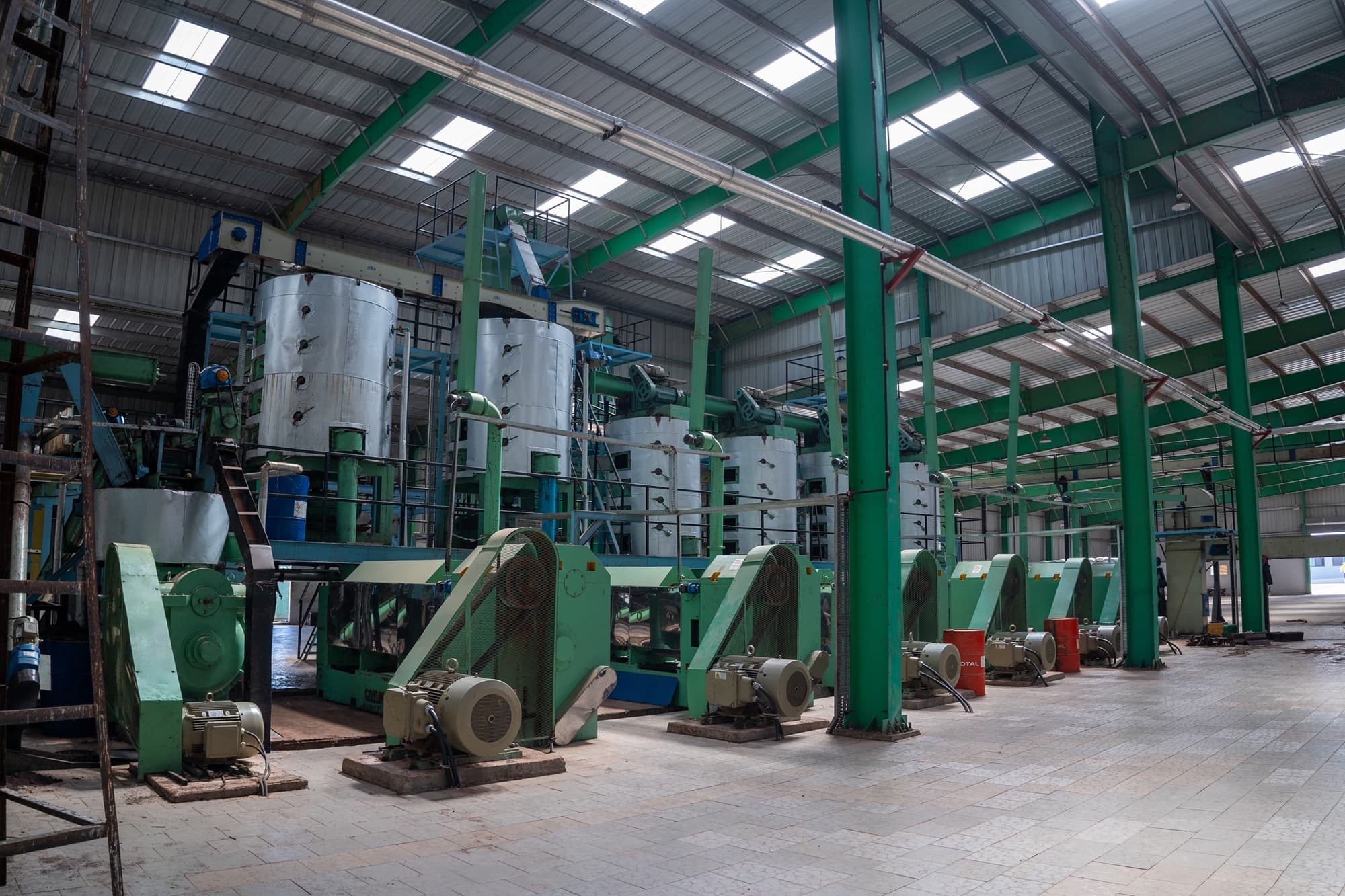 Intérieur d’une usine de transformation agroalimentaire : longue rangée de presses vertes à moteur électrique, cuves inox et convoyeurs sous une charpente métallique à verrières. — API-Mali