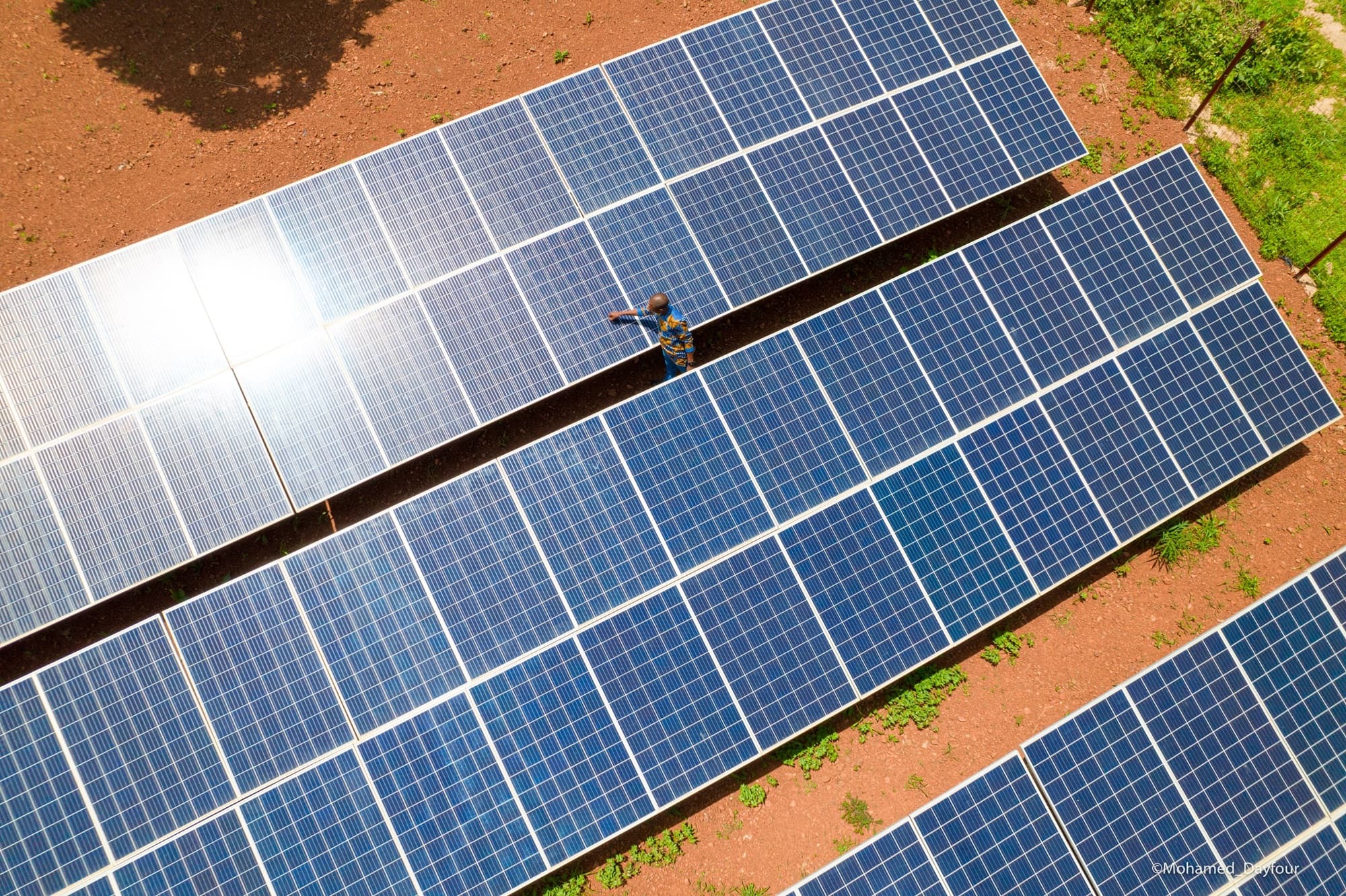 Vue en plongée sur trois longues rangées de panneaux photovoltaïques bleus posés sur un sol de latérite rouge, un technicien nettoyant un panneau entre deux rangées. — Mohamed Dayfour / API-Mali