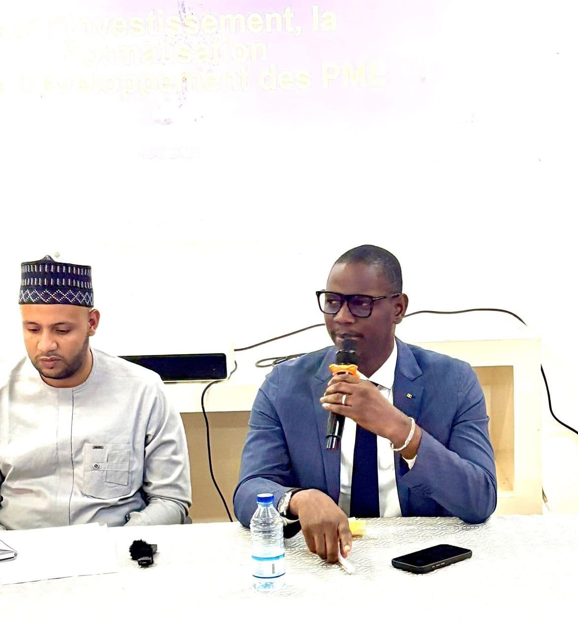 Deux hommes assis à une table d’honneur nappée de blanc sous une diapositive projetée, celui de droite parlant dans un micro à main, celui de gauche lisant ses notes. — API-Mali